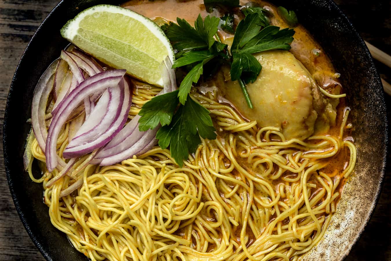 khao soi, thai curry noodle in a black bowl with lime wedge and chicken.