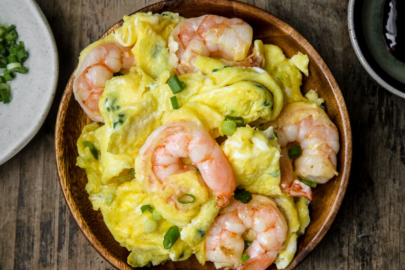 A plate of Chinese scrambled eggs with shrimp in a wooden plate served with green onions on top.