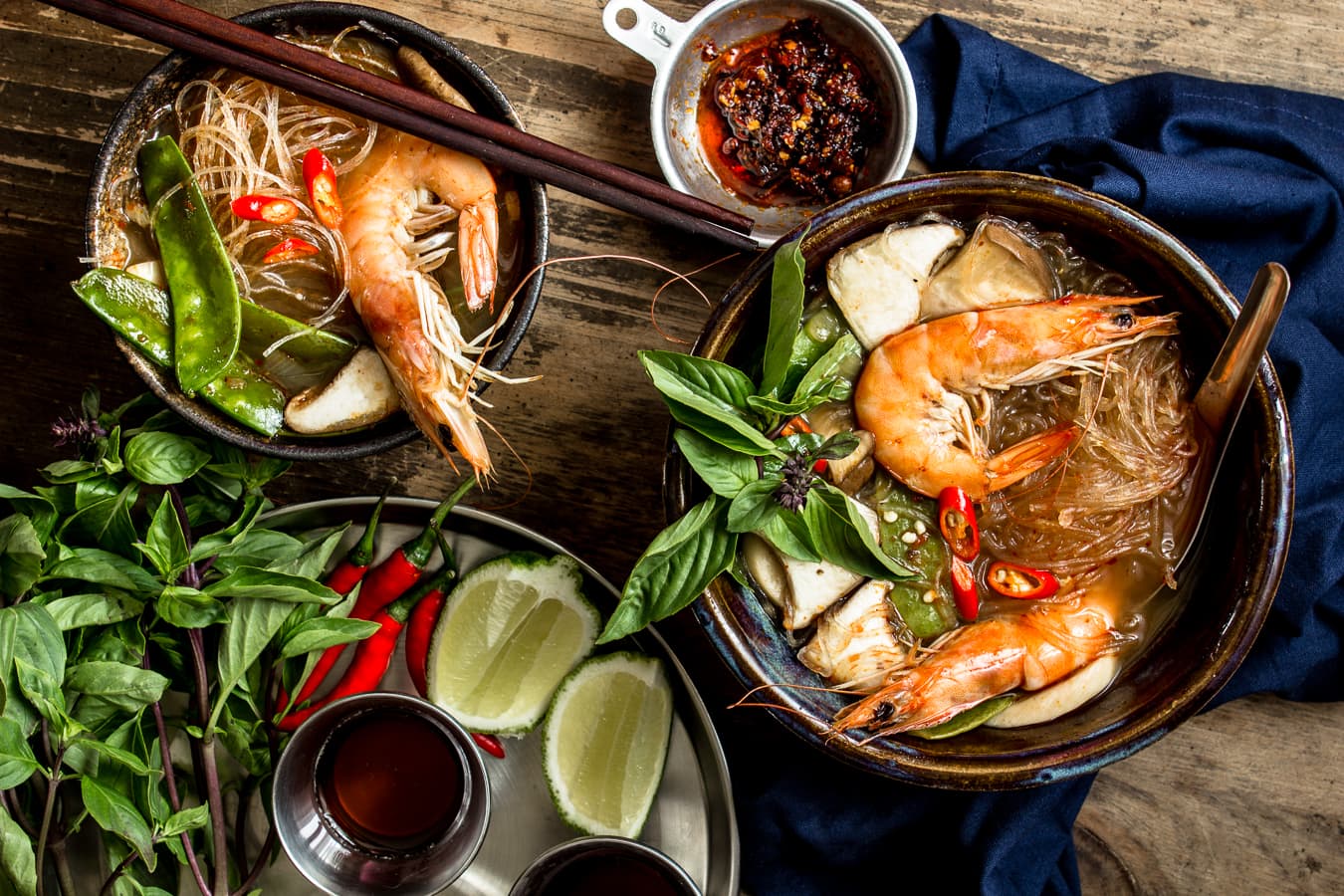 Two bowls of tom yum goong served with shrimp and glass noodles in brown bowls on a wooden table