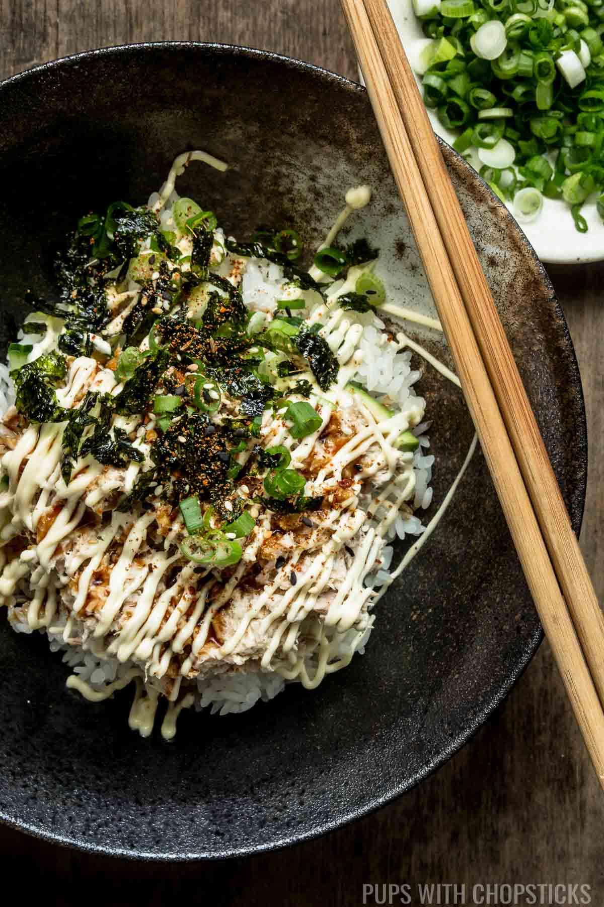 A bowl of rice with mayo tuna on top garnished with green onions and seaweed, served in a black bowl