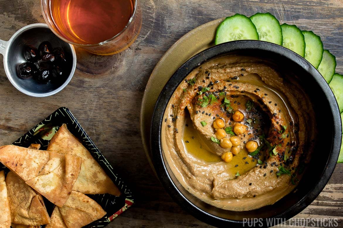 Black garlic hummus in a bowl served with cucumbers, pita chips and black olives on a wood table