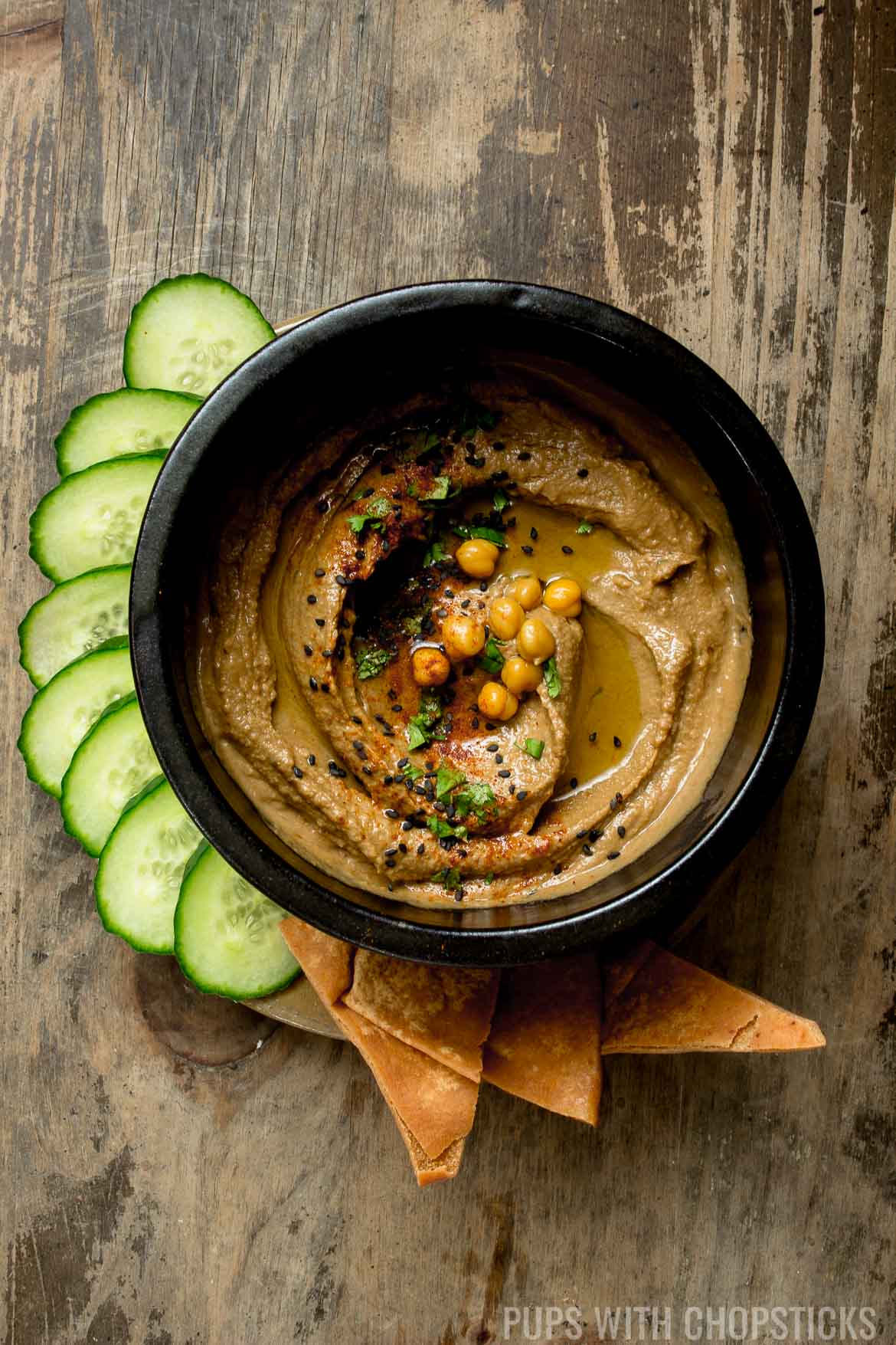 Black garlic hummus in a large black bowl served with cucumbers and pita chips on a wooden table