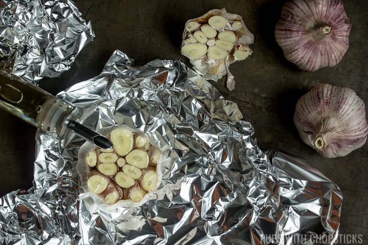 Garlic bulb with top cut off, being drizzled with oil and wrapped in foil in preparation for roasting in the oven