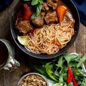 Bo Kho served with noodles, basil, lime wedges on a wooden table with a cup tea