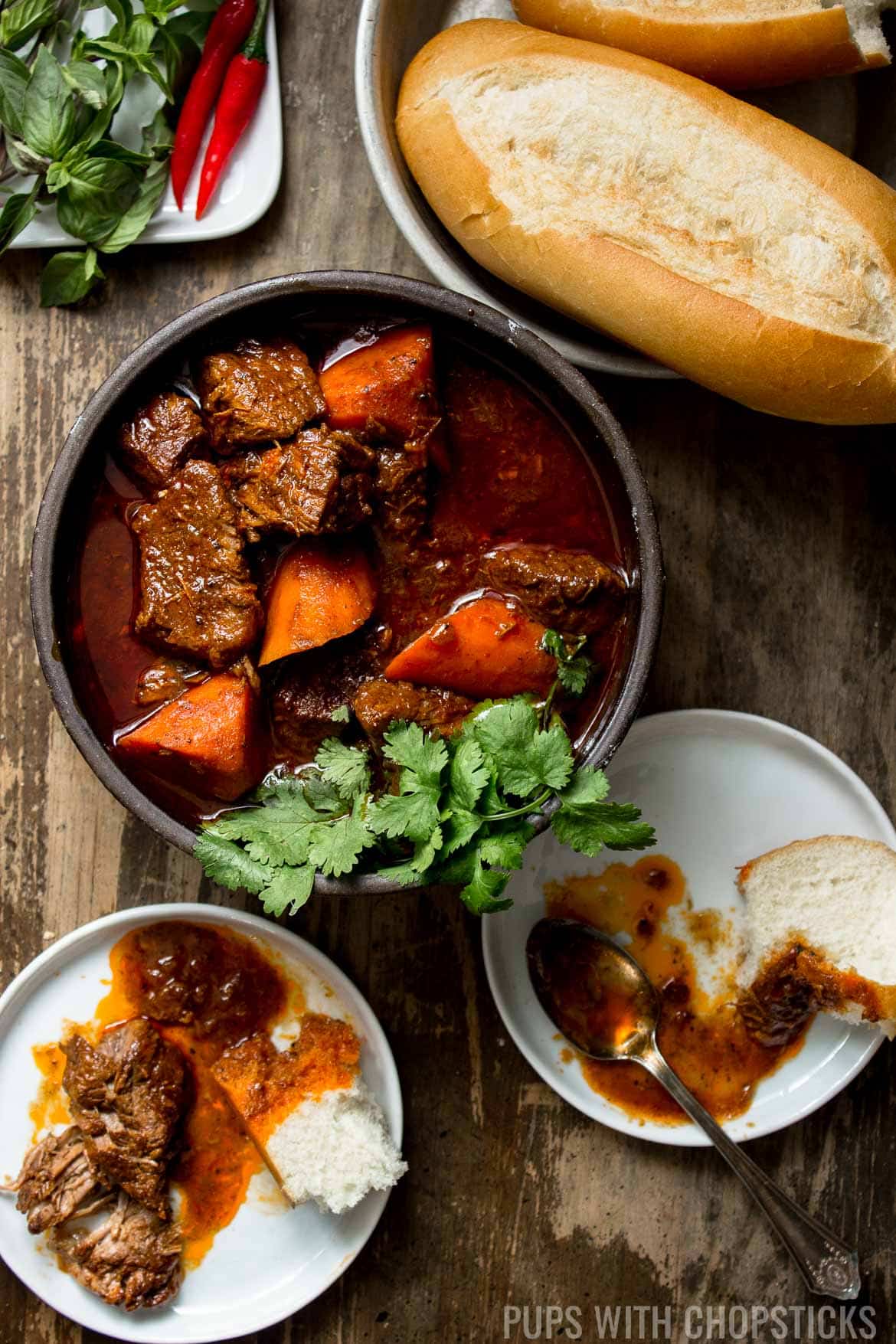 A large bowl of bo kho on a wooden table, being eaten with ripped pieces of Vietnamese baguette