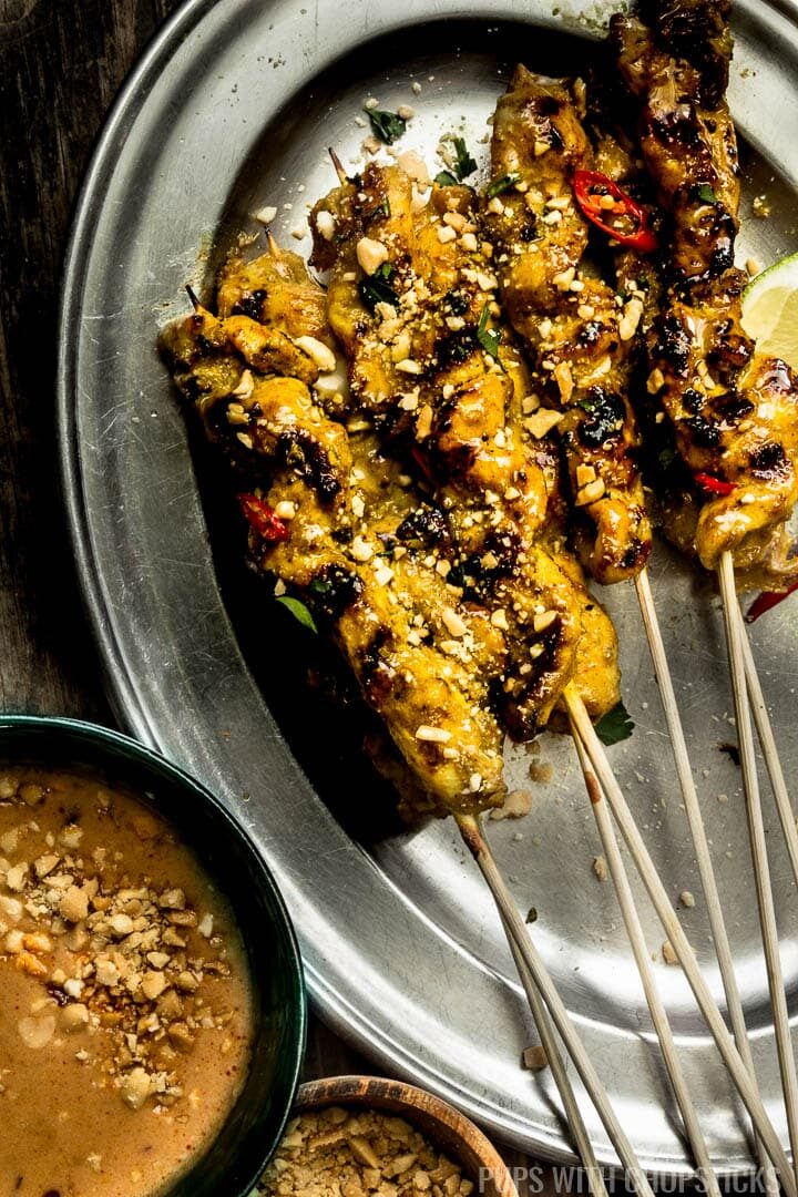 Multiple Thai chicken satay skewers on a metal plate with a bowl of peanut sauce on the side.