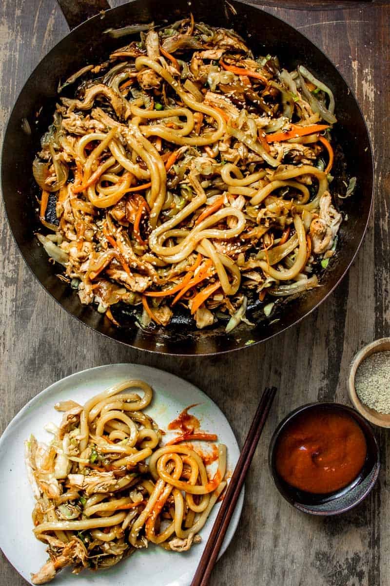 A pan with Stir Fry Chicken Teriyaki yaki udon noodle stir fry, being served on a plate with hot sauce and sesame seeds