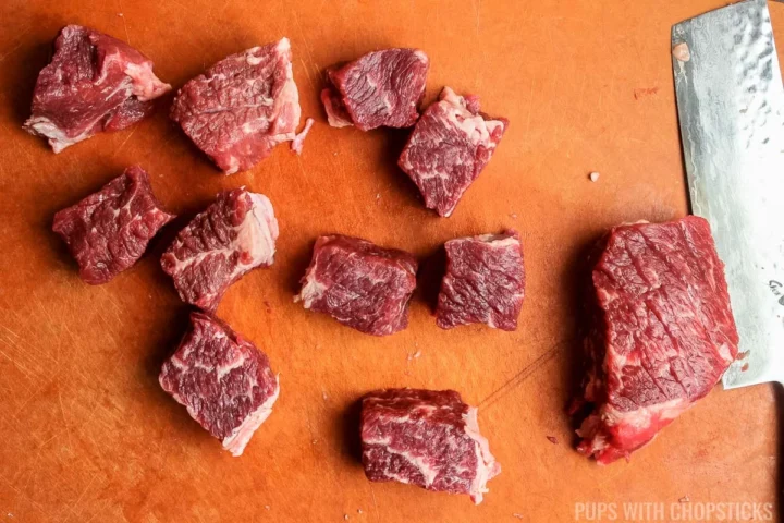 rib eye steak being cut into 1 inch cubes on a cutting board.