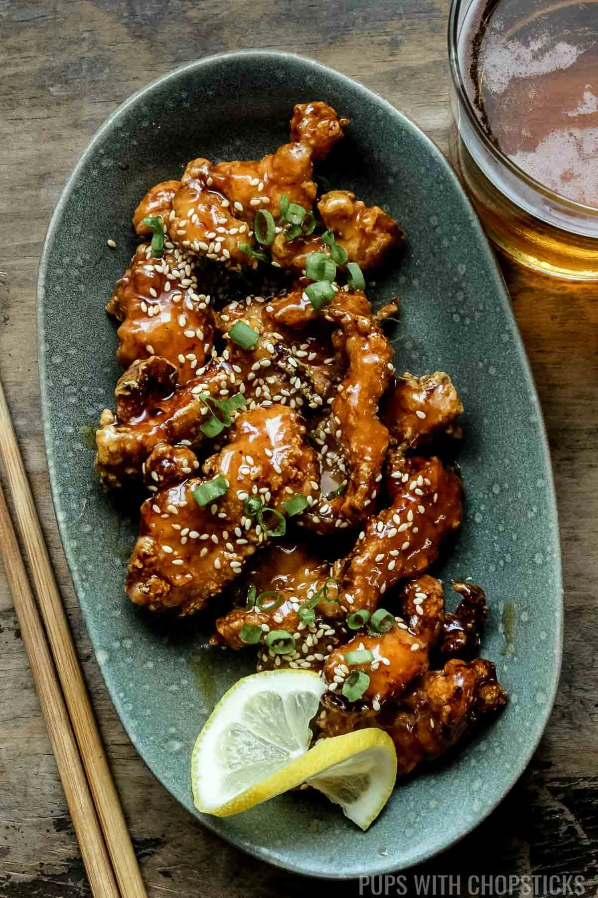 Bite-sized crispy Chinese lemon chicken tossed in a lemon sauce and served on a green plate with a lemon slice with a glass of beer and wooden chopsticks.