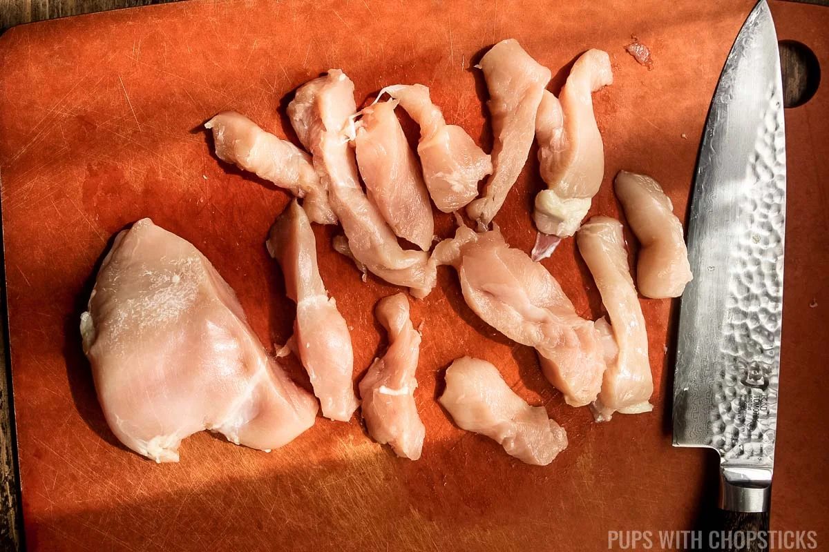 Raw chicken chunks being cut on a cutting board.
