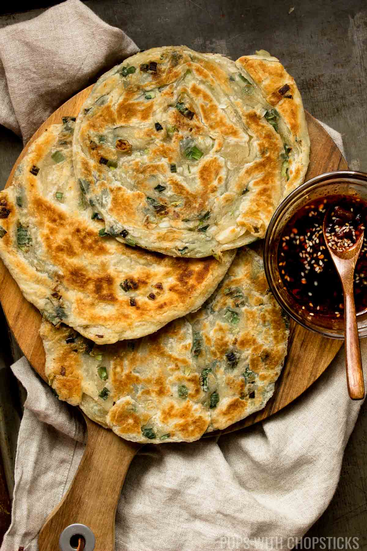3 scallion pancakes on a wooden board served with a side of dipping sauce.