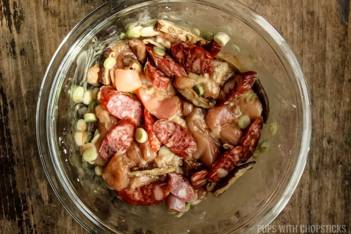 A bowl of marinating chicken, chinese sausage, green onions and shiitake mushrooms in a glass bowl.