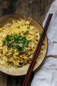 Large bowl of egg fried rice topped with green onions and sesame seeds with chopsticks and handkerchief on wooden table.