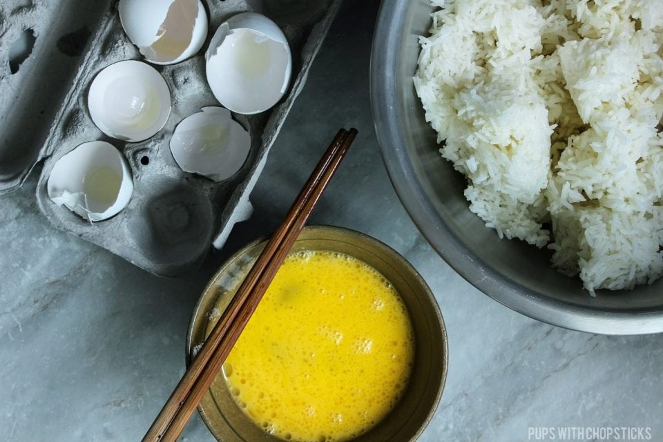 Small bowl of raw beaten eggs with chopsticks and with a bowl of day old rice with egg shells in a carton