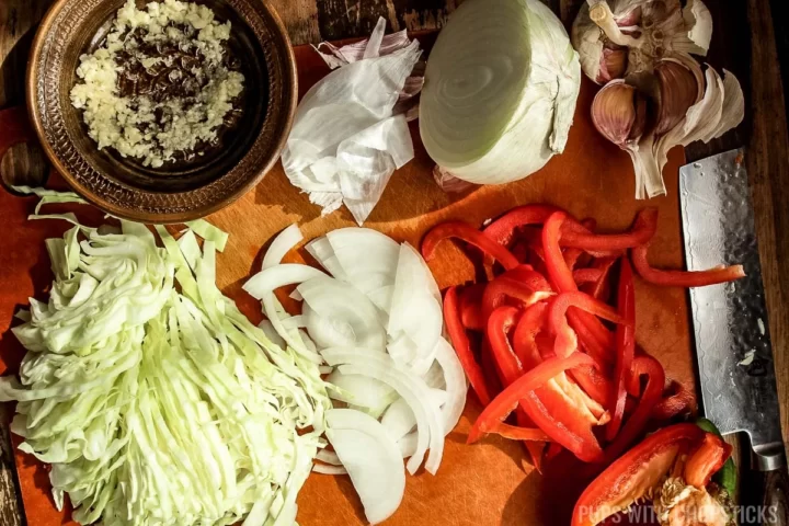 A cutting board with chopped cabbage, red bell pepper, white onion and grated garlic.