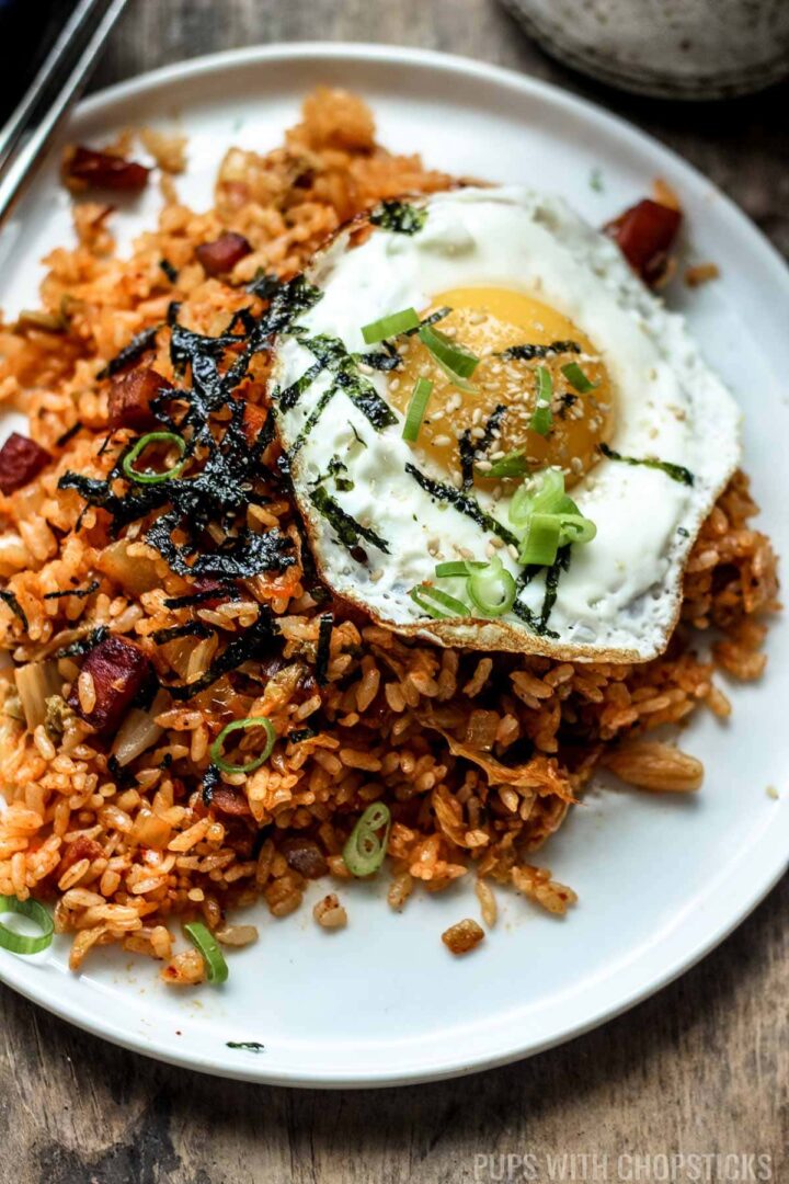 A small white plate of Korean kimchi fried rice topped with a fried sunny side up egg and garnished with green onions, sesame seeds and seaweed.