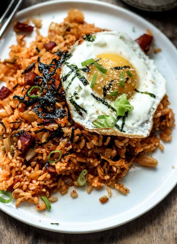 A small white plate of Korean kimchi fried rice topped with a fried sunny side up egg and garnished with green onions, sesame seeds and seaweed.