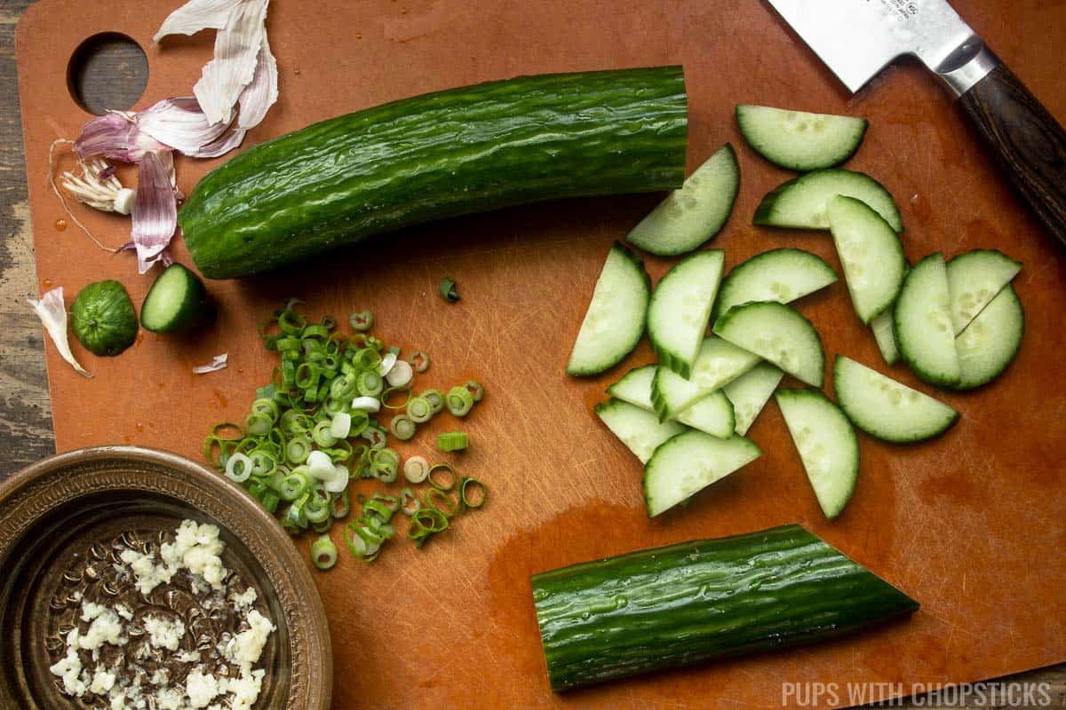 A cutting board with sliced cucumbers, chopped green onions and grated garlic.