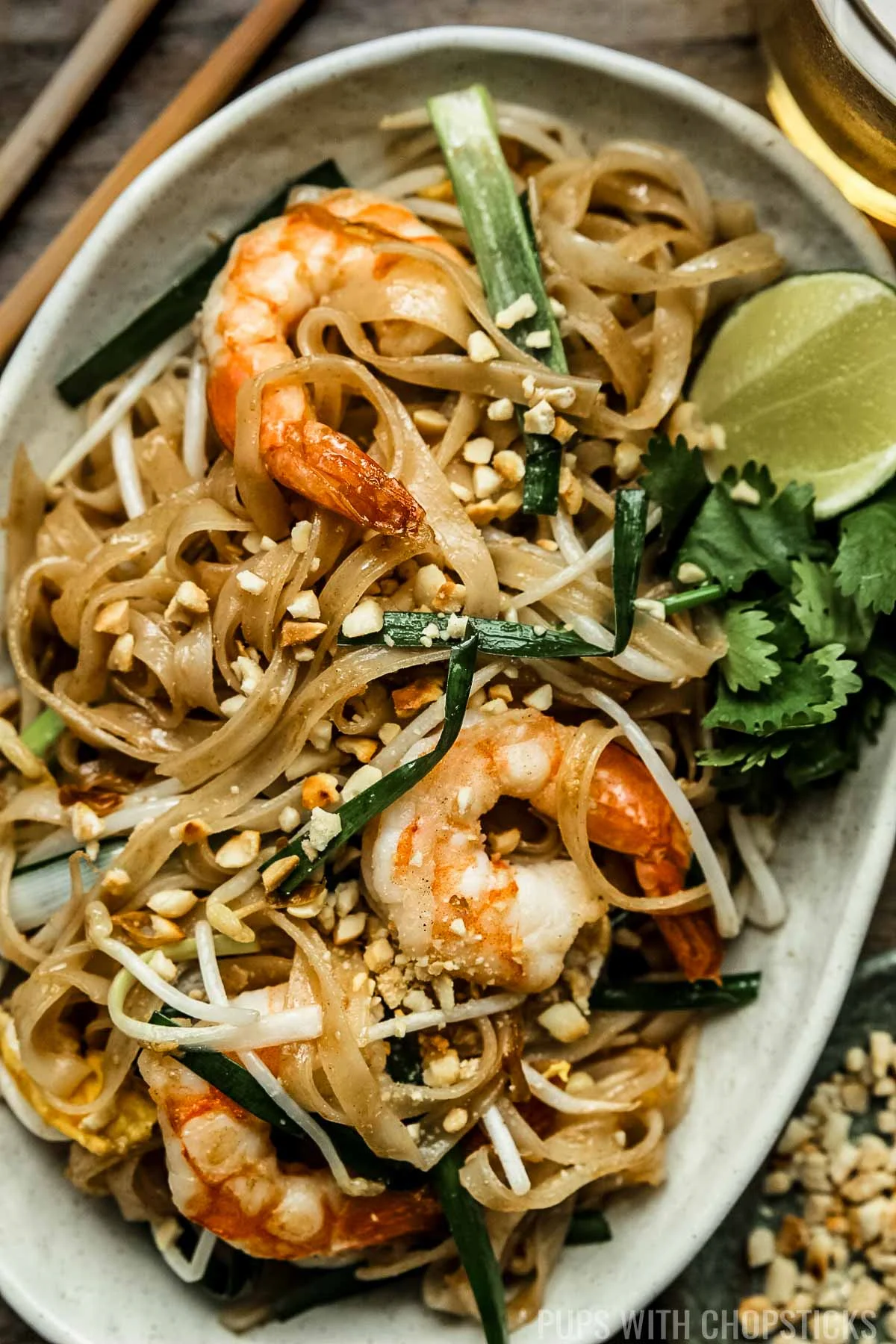 Pad thai with shrimp on a beige oval plate served with crushed peanuts and lime wedges