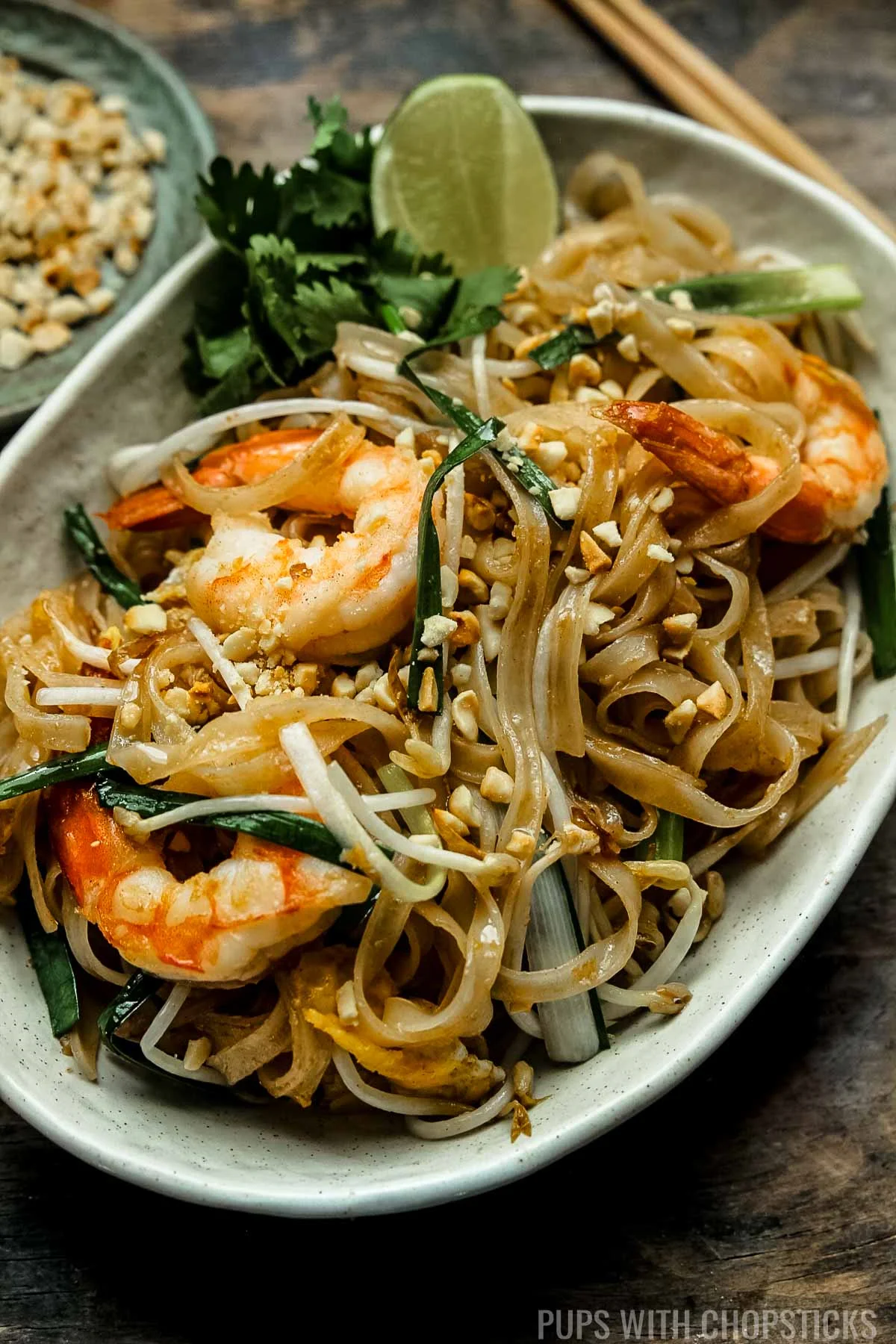 Angled photograph of pad thai with shrimp, on a wooden table with chopsticks.