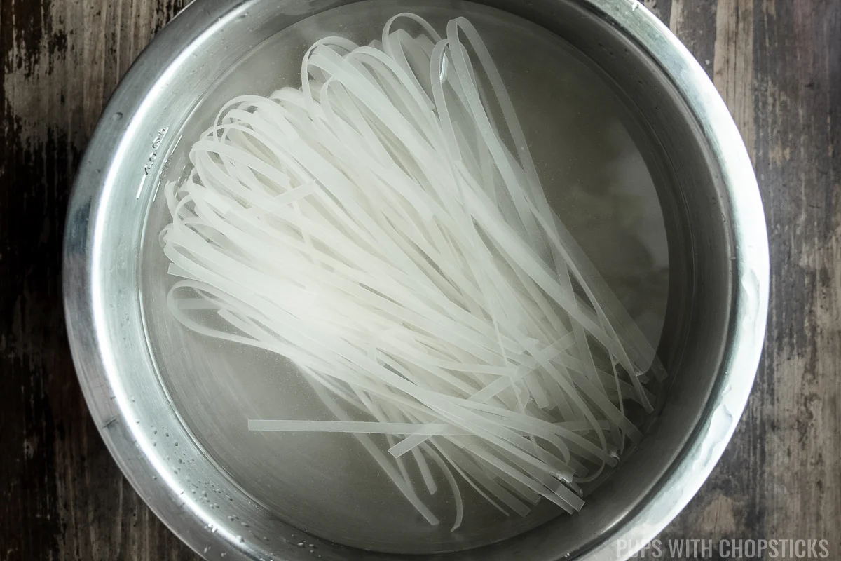 Rice noodles being soaked in hot water in a metal bowl in prep for pad thai