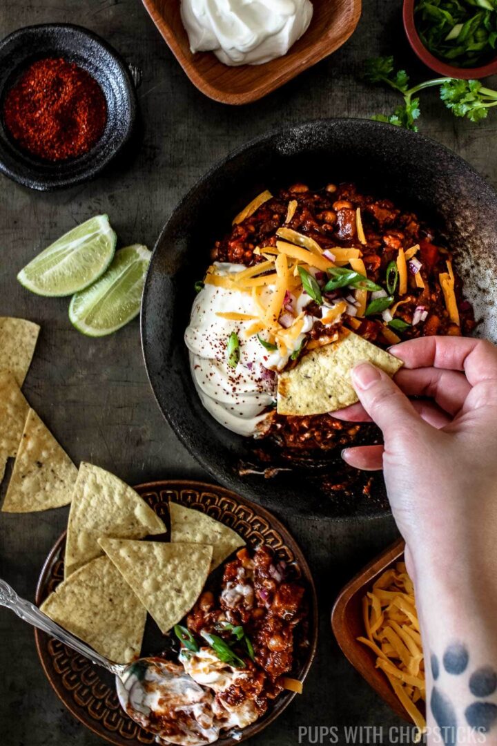 Sweet and Smoky Pineapple Chipotle Chili in a bowl with chips, lime wedges, sour cream and cheese on a table being scooped with chips
