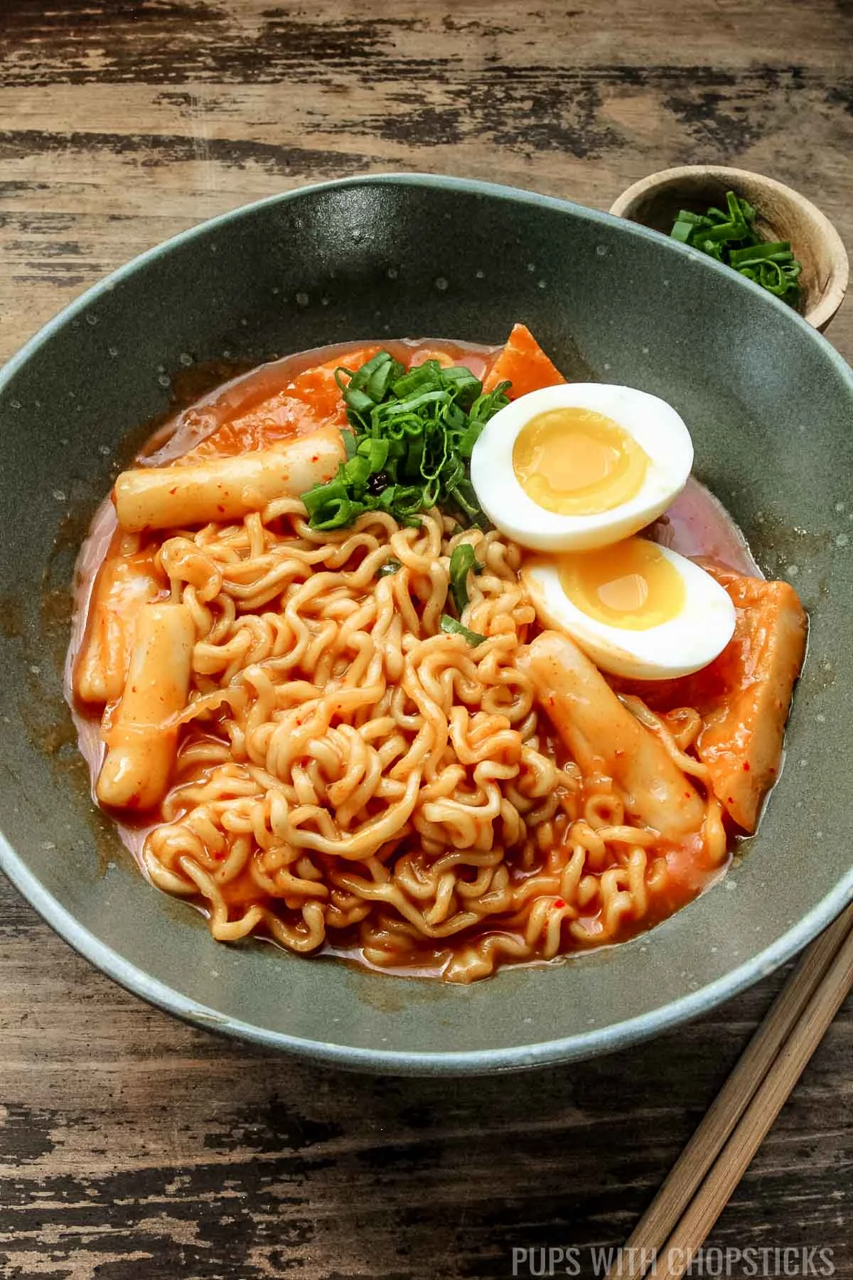 Closeup of Rabokki served with a soft boiled egg in a green bowl with chopsticks.