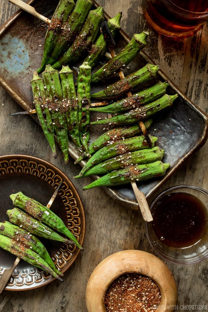 Skewered roasted okra on a plate served with a drink and a bowl of extra spices on the side