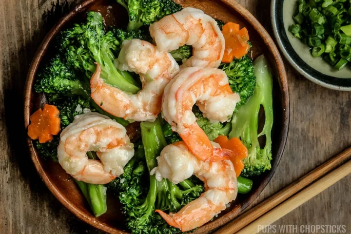 Shrimp and broccoli in a brown plate with a side of green onions and chopsticks.