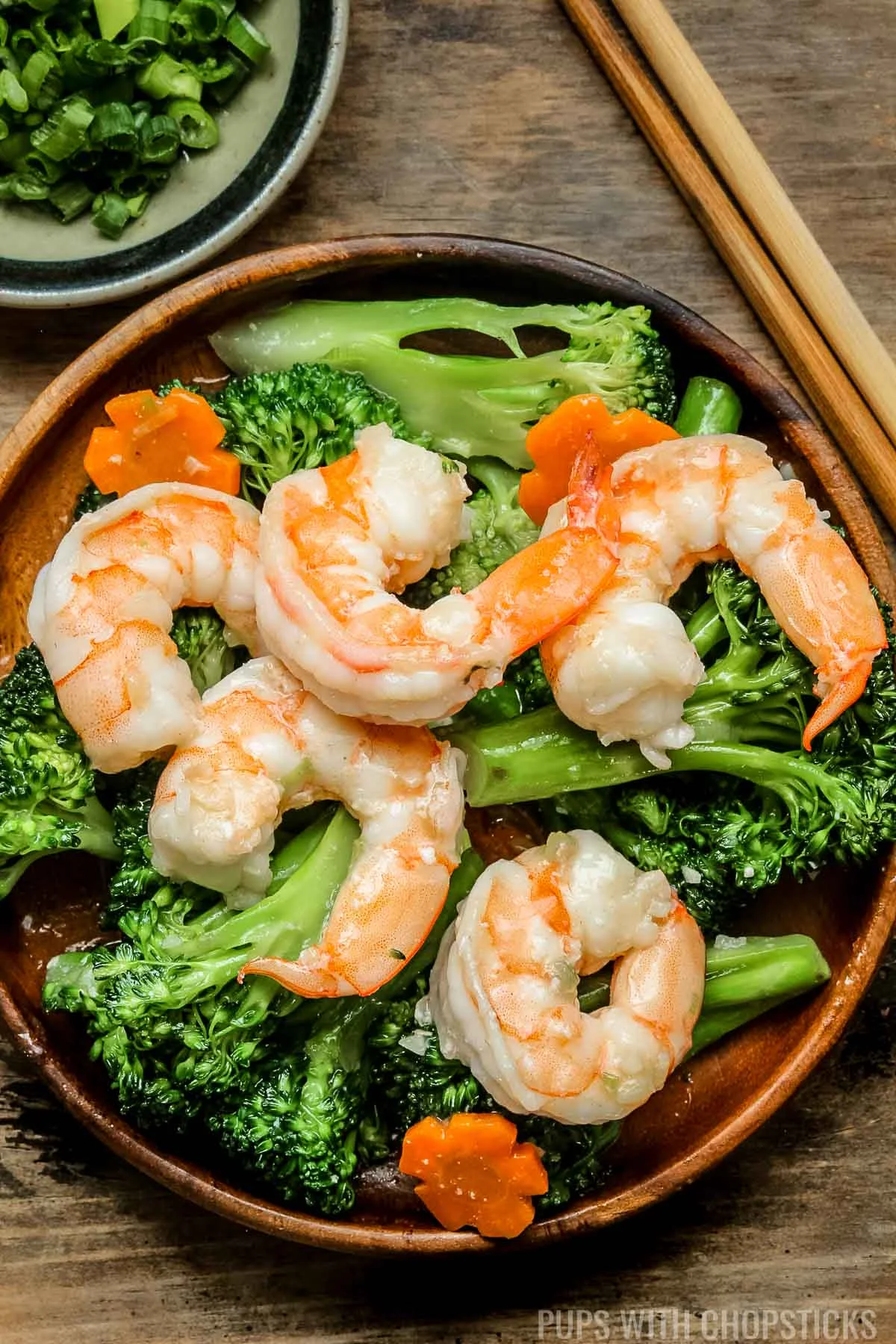 Shrimp and broccoli in a brown plate with a side of green onions and chopsticks.