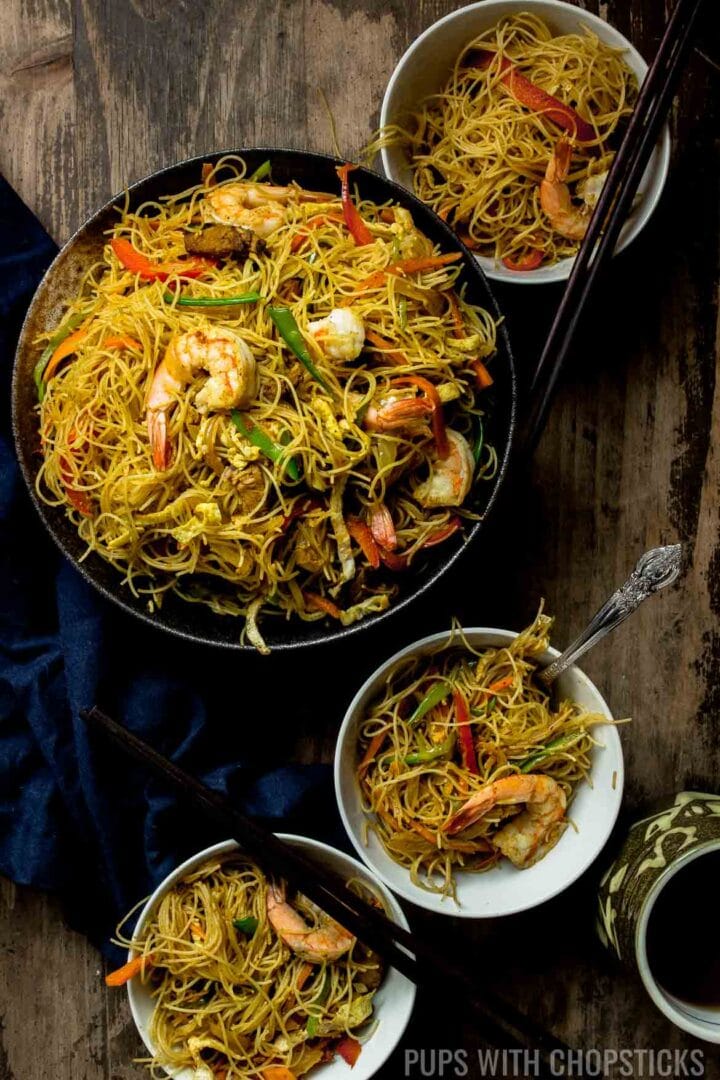 A large bowl of Singapore noodles, with shrimp, red pepper, snow peas, eggs and dry curried noodles, served into 3 small bowls