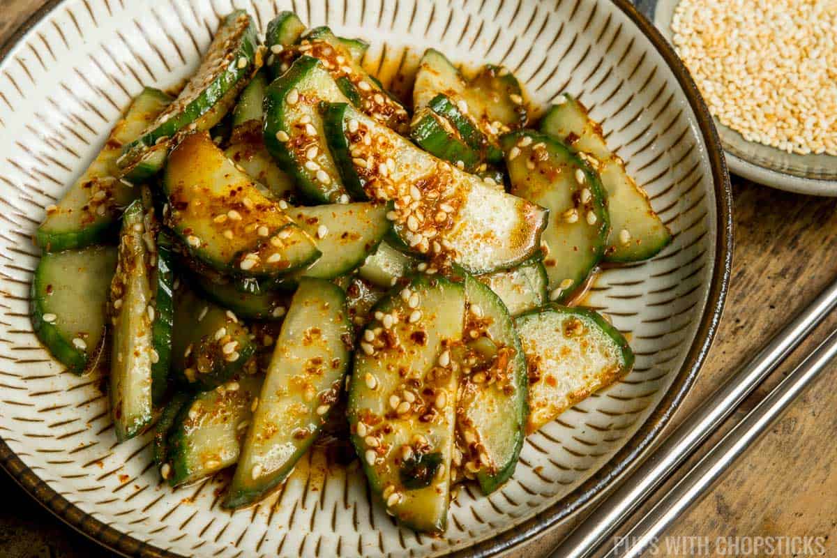 Close up of Korean cucumber salad (oi muchim) served with a side of toasted sesame seeds with metal chopsticks.
