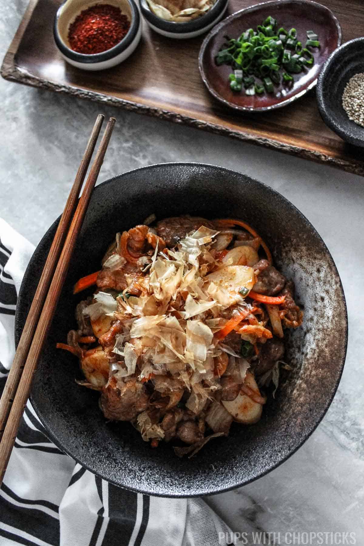 Stir-fried rice cakes with bonito flakes on top served in a black bowl.