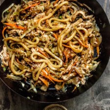 A large frying pan with chicken teriyaki yaki udon inside, with small bowls of sesame seeds and hot sauce on the side.
