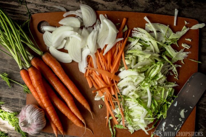 Carrots, onions, cabbage and garlic being sliced on a cutting board in preparation for Stir Fry Chicken Teriyaki Noodles
