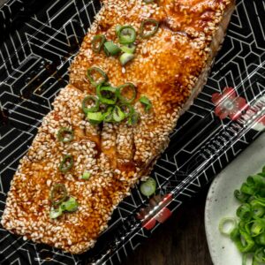 A salmon filet crusted with sesame seeds with teriyaki sauce on a takeout tray on a wooden table