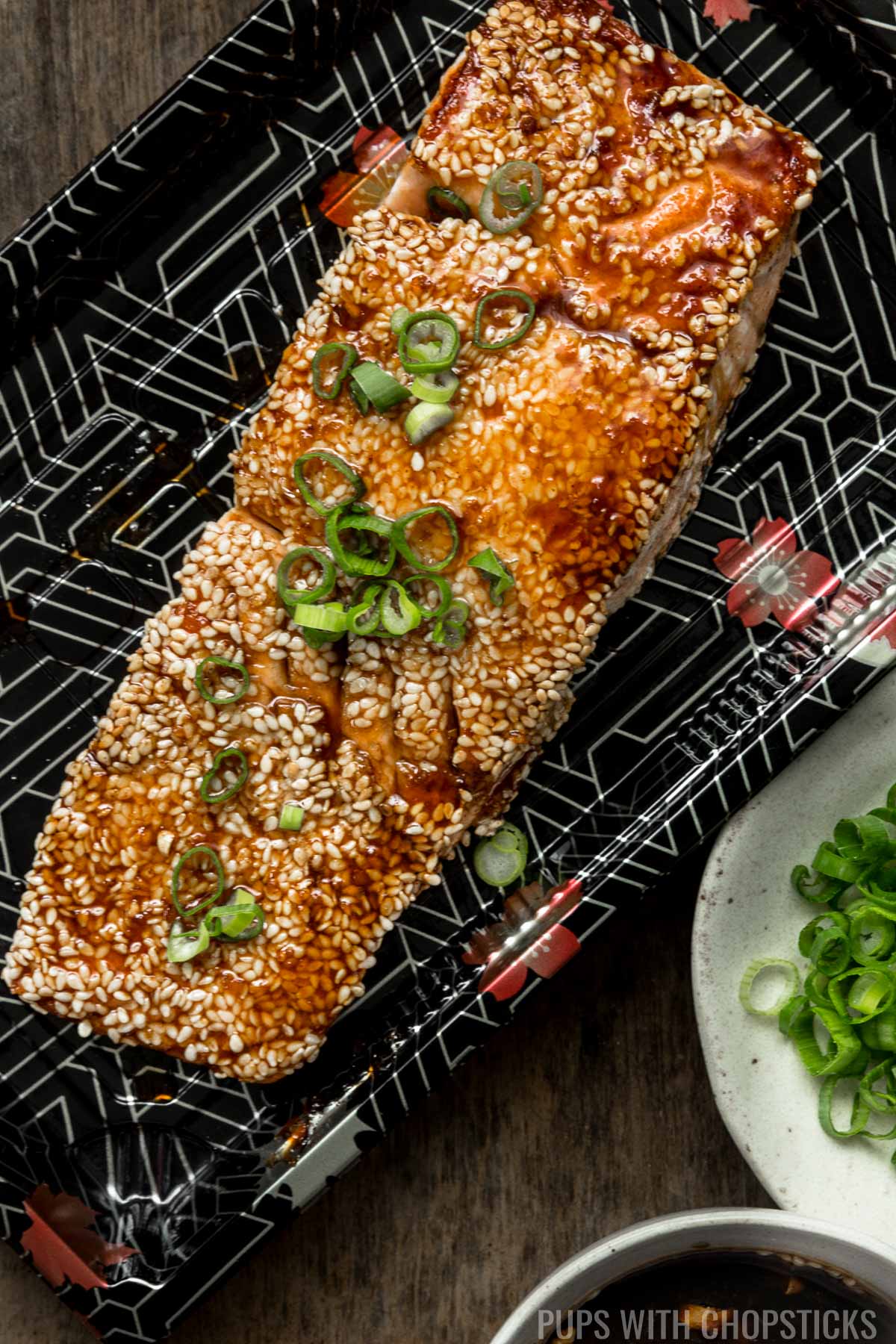 A cooked salmon filet, crusted with sesame seeds and glazed with teriyaki sauce in a takeout plate on a wooden table