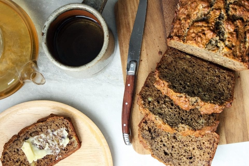 A slice of banana bread being buttered and served with tea