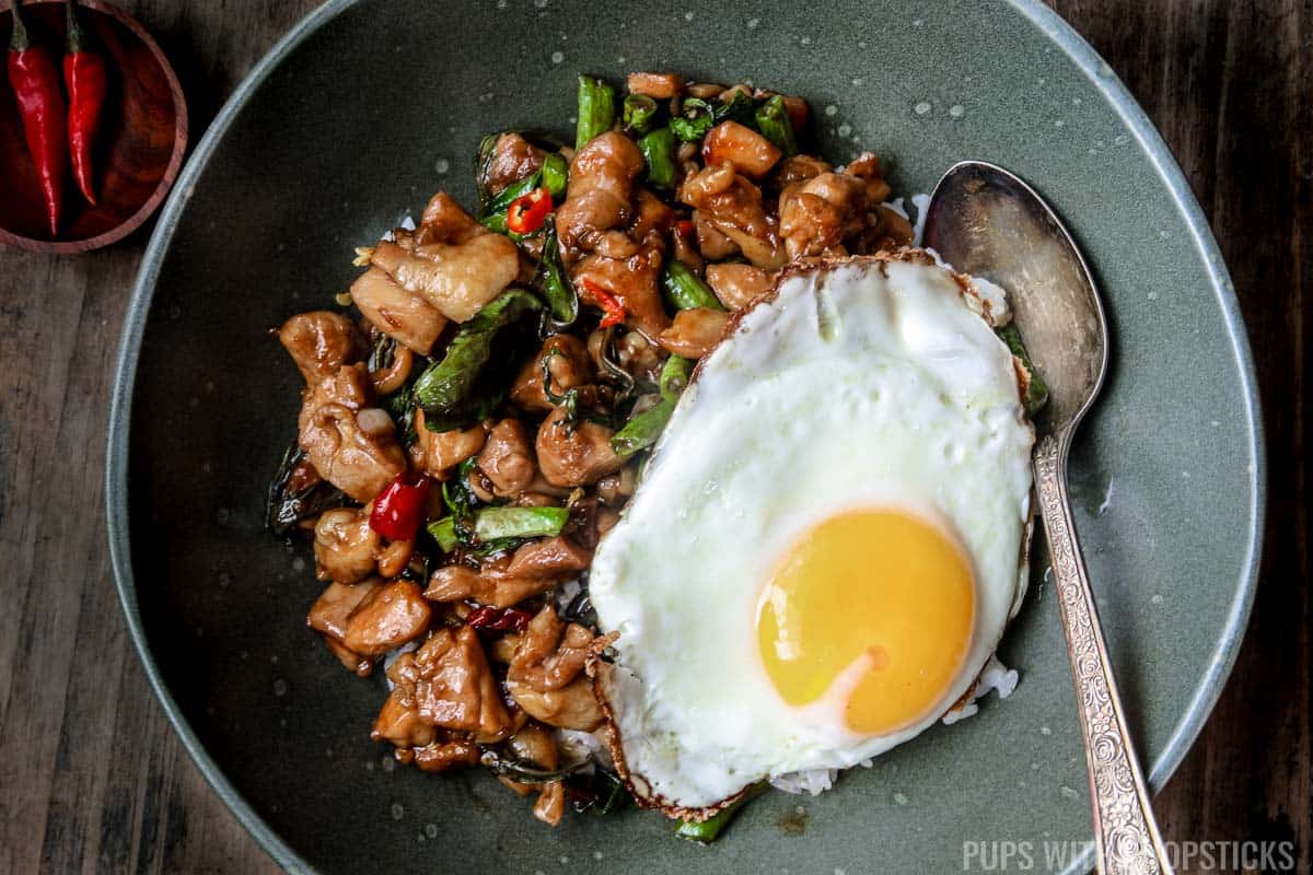Thai basil chicken being served over jasmine rice with a fried egg in a green bowl.