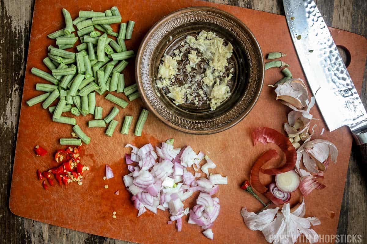A cutting board with minced garlic, green beans cut into ½ inch pieces, shallots diced finely, and birdseye chili minced.