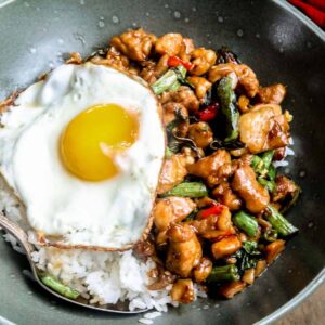 close up of thai basil chicken (pad kaprao) in a green bowl served over rice with a fried egg