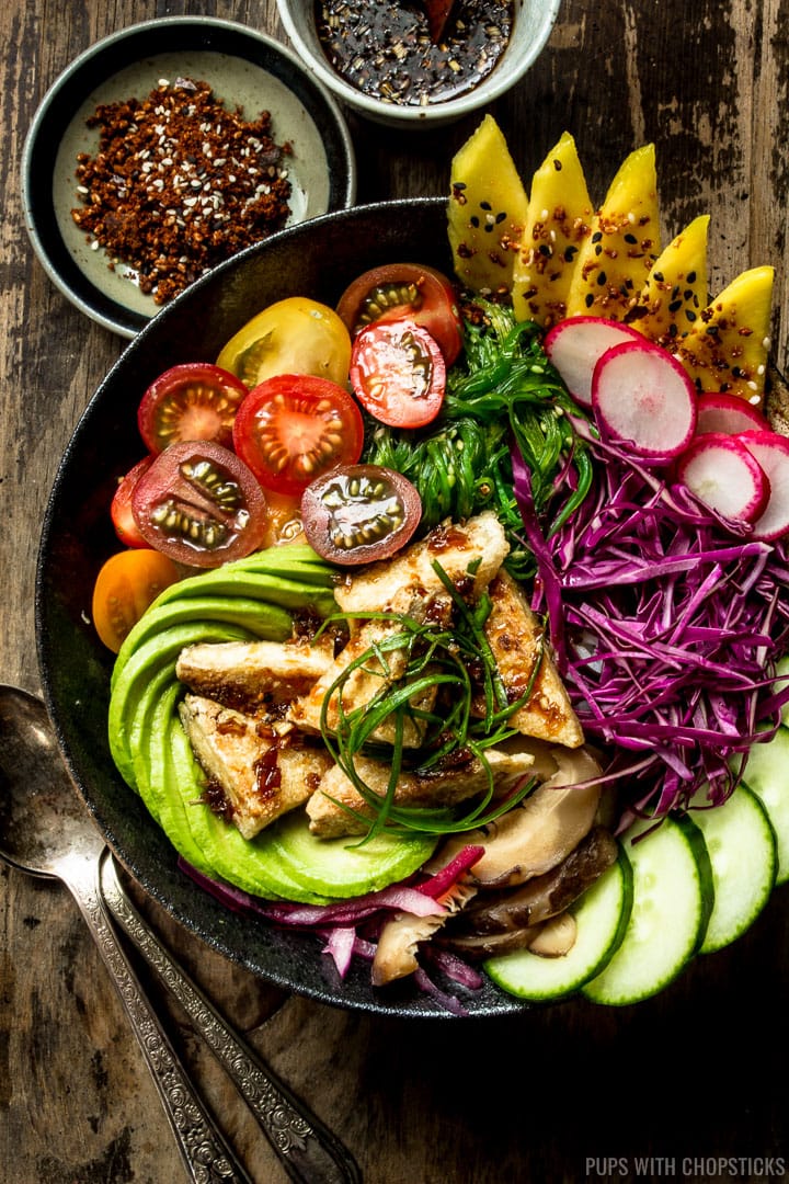 A buddha bowl filled with fresh local produce in a dark bowl with some spoons on a wooden table