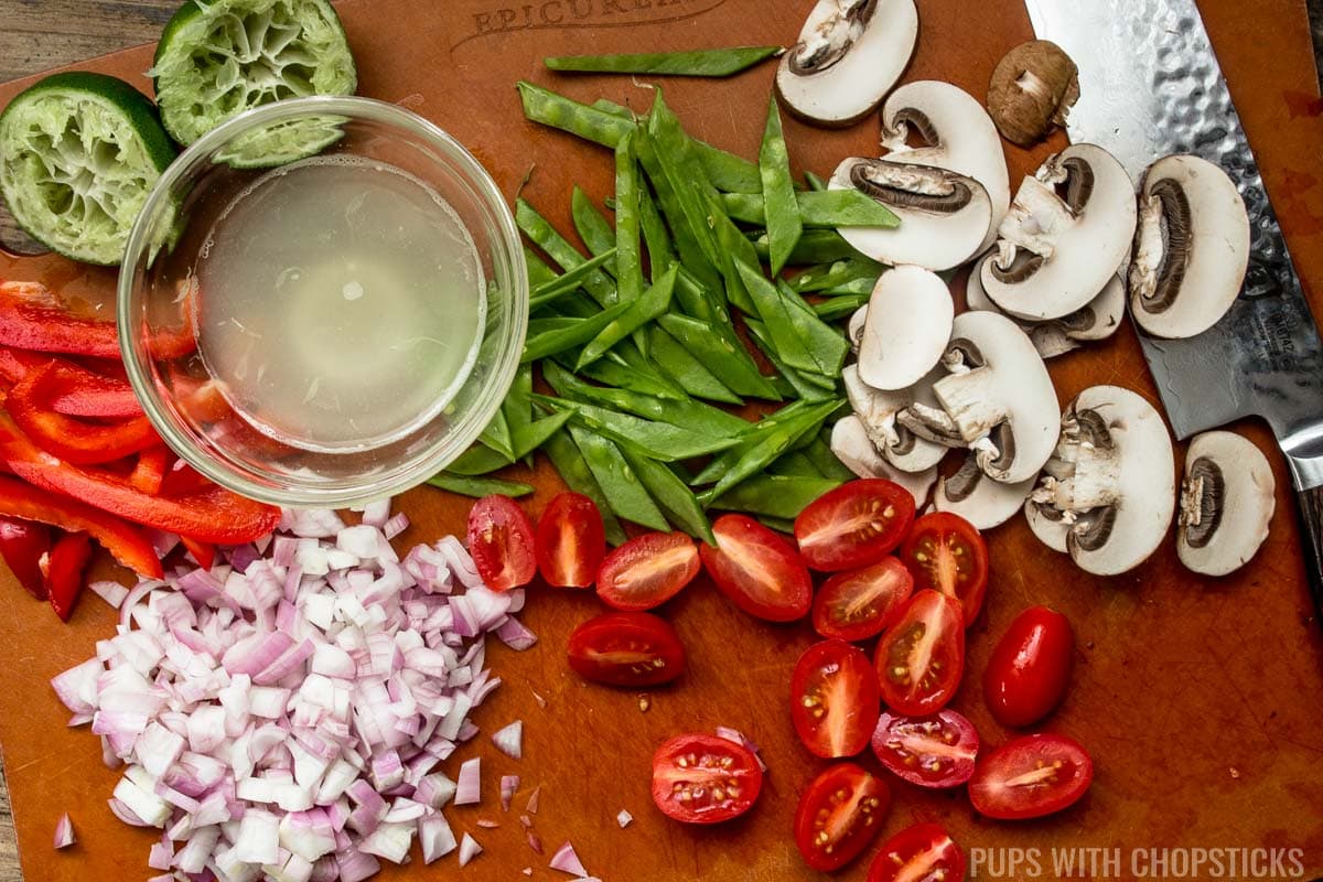 Vegetables being sliced on a cutting board (thinly sliced mushrooms, tomatoes, snow peas, shallots, sweet red peppers