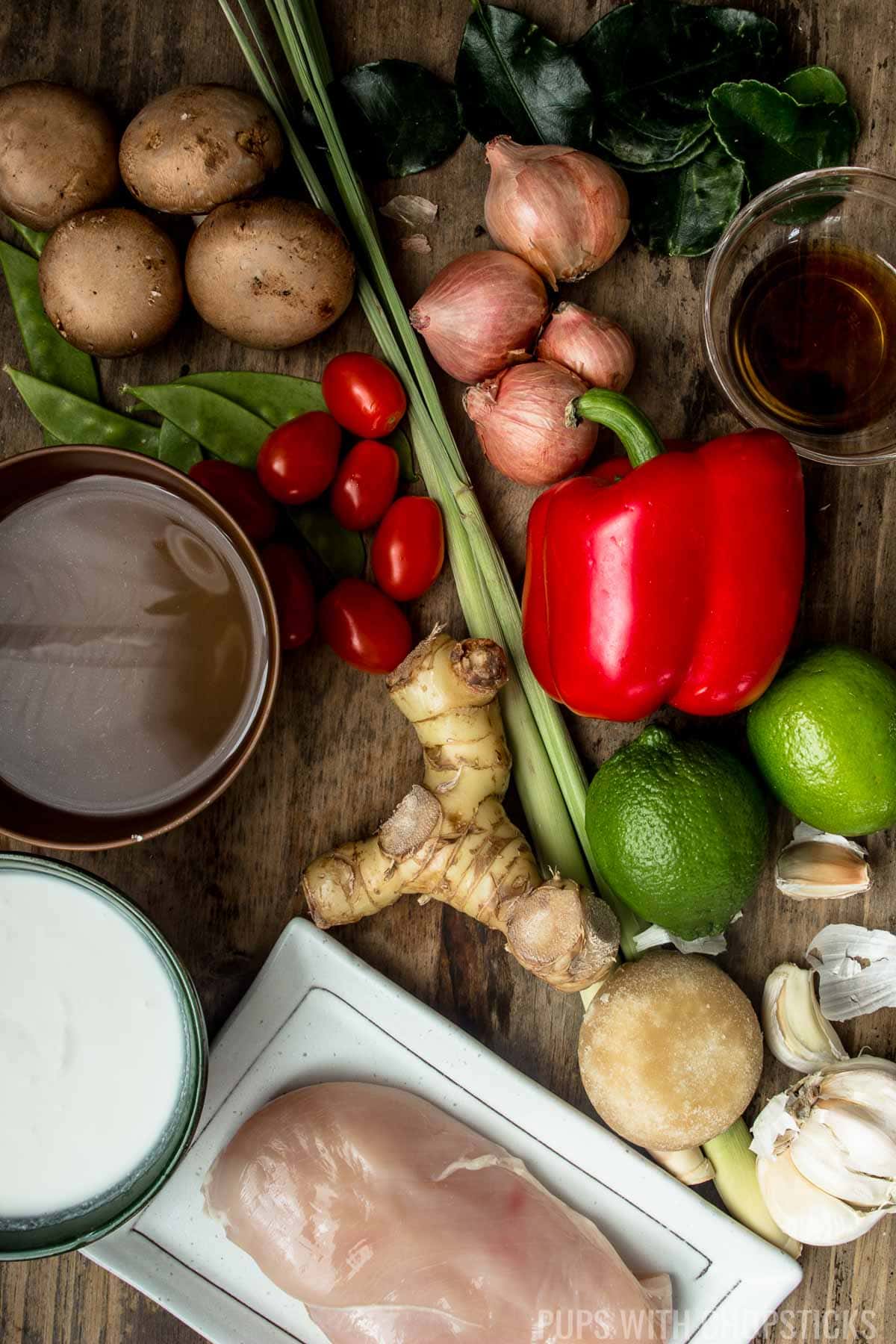 Mise en place of tom kha gai ingredients (chicken, coconut milk, broth, lemongrass, kaffir lime leaves, garlic, galangal, lime, sugar, fish sauce, onions, tomatoes, mushrooms)