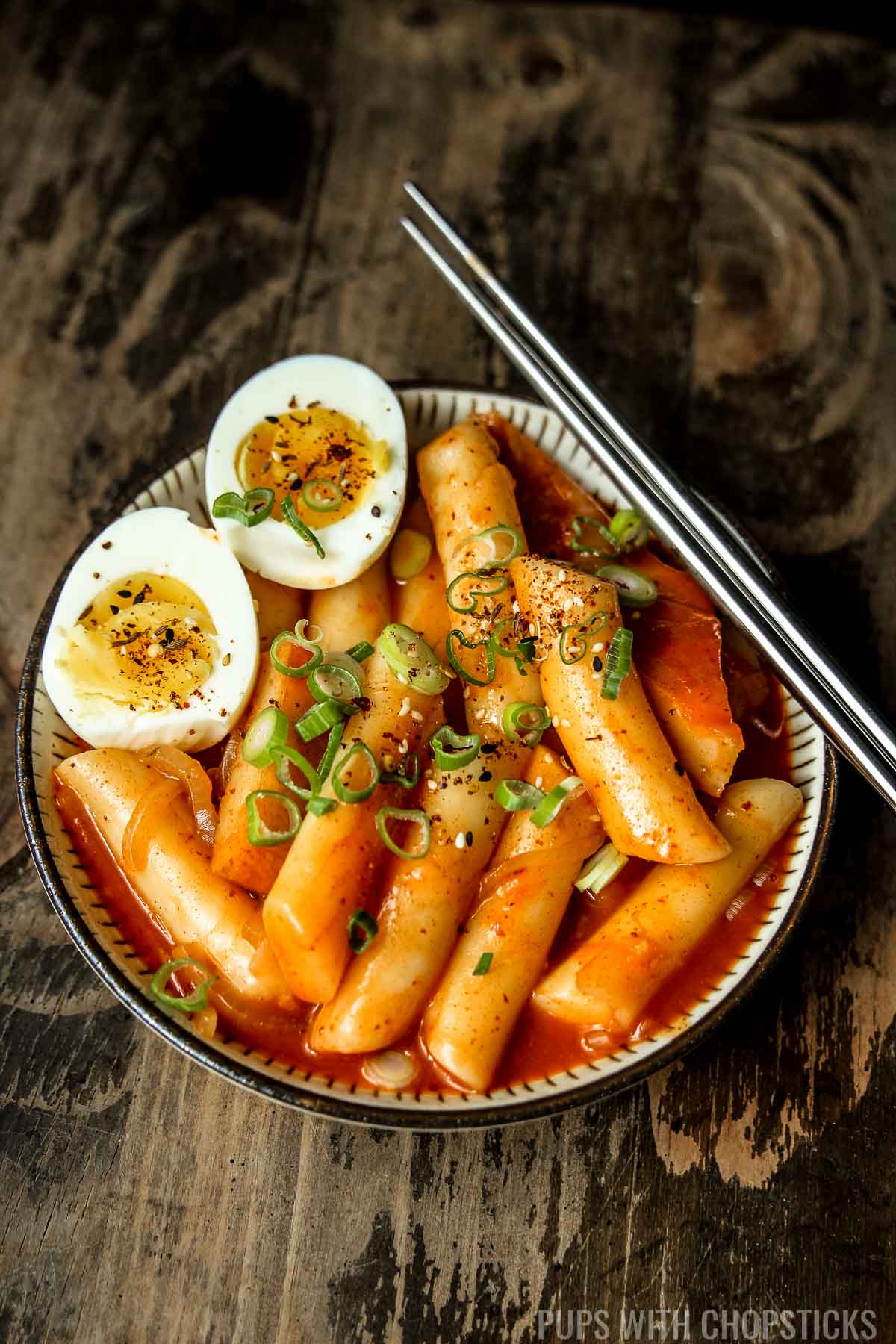 A small bowl of tteokbokki (Spicy korean rice cakes) with medium boiled eggs sliced in half and garnished with green onions, with a pair of metal chopsticks