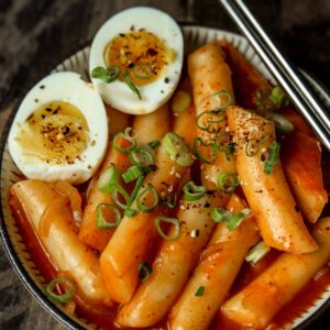 A small bowl of tteokbokki (spicy korean rice cakes) and some medium boiled eggs sliced in half and served with metal chopsticks w