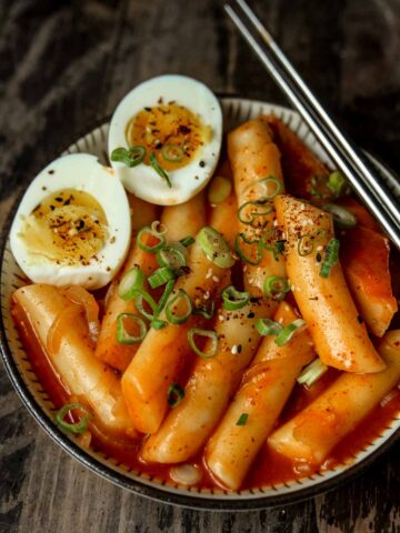 A small bowl of tteokbokki (spicy korean rice cakes) and some medium boiled eggs sliced in half and served with metal chopsticks w