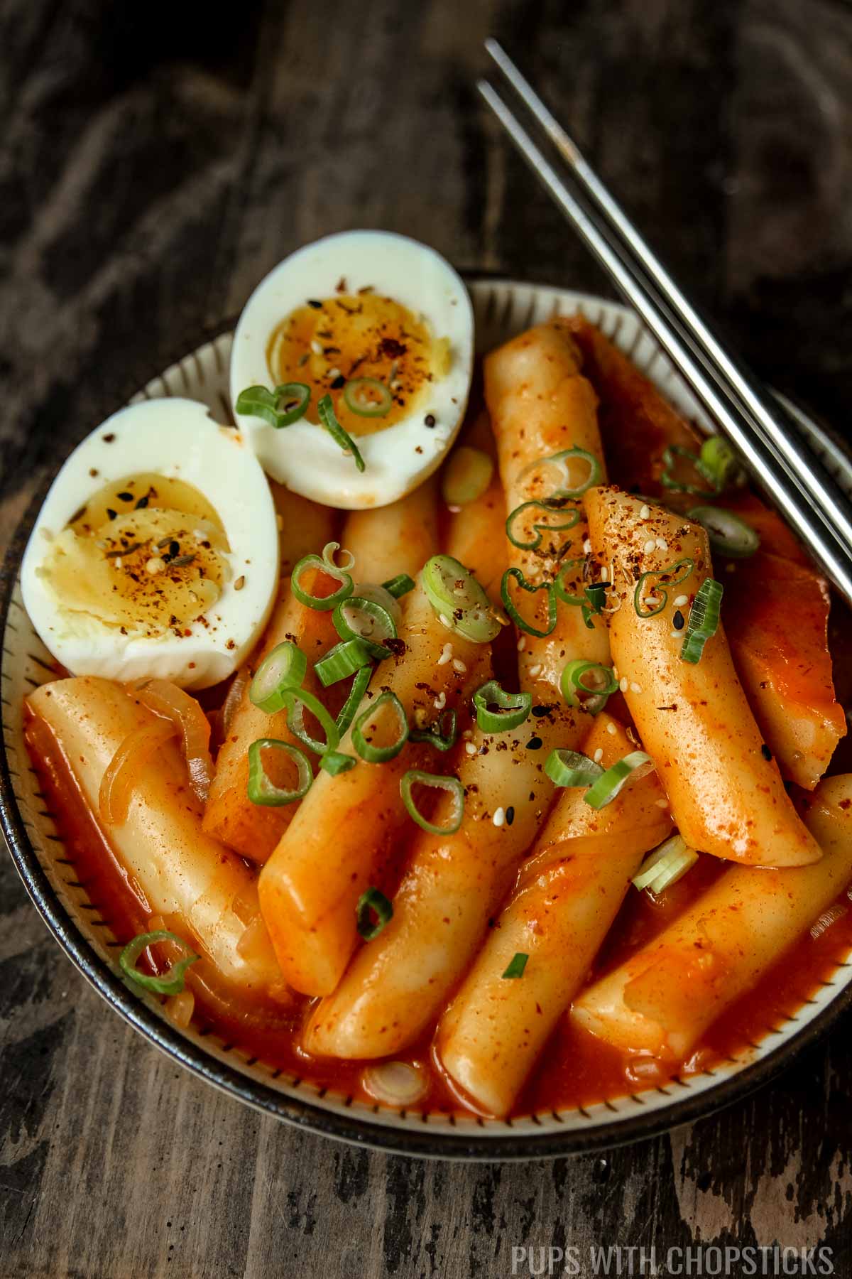 Closeup of tteokbokki with soft boiled eggs and metal chopsticks on a wooden table.