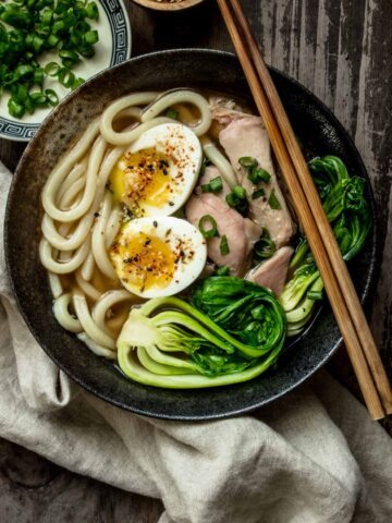A large bowl of turkey udon noodle soup with soft boil egg, leftover turkey and bok choy on top, with green onions on the side.