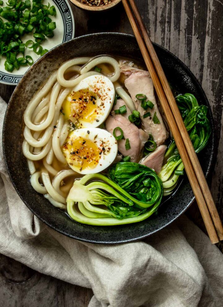 A large bowl of turkey udon noodle soup with soft boil egg, leftover turkey and bok choy on top, with green onions on the side.